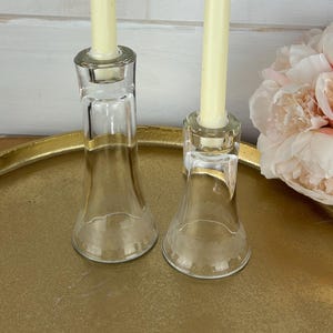 May include: Two clear glass candle holders with ivory taper candles. The candle holders are on a gold tray, with a bouquet of pink flowers in the background. The candle holders have a flared base.