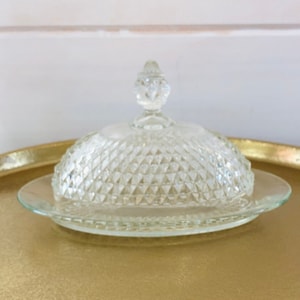 May include: A clear glass butter dish with a diamond pattern and a knob on the lid. The dish is sitting on a gold tray.
