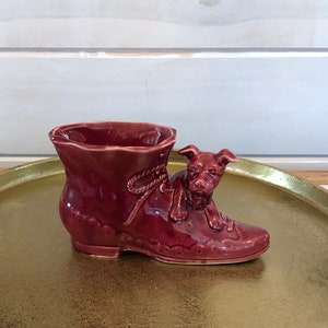 May include: A red ceramic planter shaped like a shoe with a dog peeking out of the top. The planter is sitting on a gold tray.