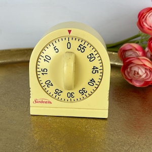 May include: A pale yellow Sunbeam kitchen timer with a rounded top and a central dial. The face features black numerical markings from 0 to 55 in five-minute increments, a red triangle indicator at the top, and the red Sunbeam brand logo at the bottom.