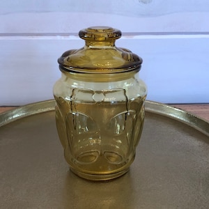May include: A clear amber glass jar with a lid, sitting on a gold tray. The jar has a decorative pattern of raised circles.
