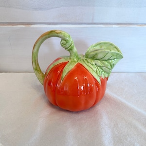 May include: Orange ceramic pumpkin pitcher with green leaves and a handle. The pitcher is sitting on a white lace tablecloth.