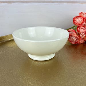May include: A small, off-white ceramic bowl with a pedestal base sits on a gold-colored tray. A bouquet of orange and pink flowers is placed to the right of the bowl. The background is a white wooden surface.
