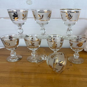 May include: A set of seven clear glass stemmed goblets featuring a frosted band around the upper rim decorated with gold leaf patterns. Each glass has a short, rounded stem and a circular base, with one glass positioned on its side in the front.