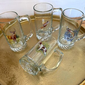 May include: Four clear glass beer mugs with handles, each featuring a different hunting-themed design. The mugs are arranged on a gold-colored tray.