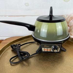 May include: An Oster electric fondue pot featuring a sage green bowl with a black lid and a long black handle. The base is black with a control dial and the text Oster Electric Fondue. A black power cord is attached to the side of the unit.