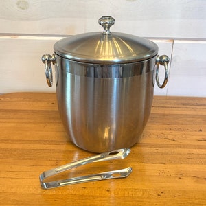 May include: A silver ice bucket with a lid and two handles. The bucket is sitting on a wooden table with a pair of silver ice tongs next to it.
