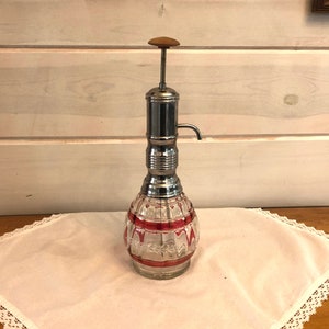 May include: A vintage glass bottle with a red and white striped design and a chrome pump dispenser. The bottle is sitting on a white tablecloth with lace trim.