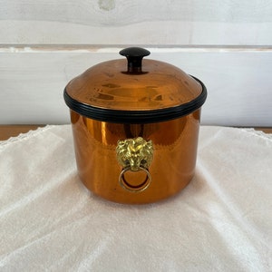 May include: A copper ice bucket with a black lid and a lion head handle. The bucket is sitting on a white lace tablecloth.