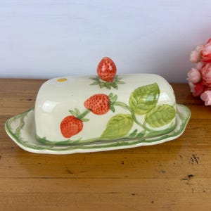 May include: A ceramic butter dish with a lid, painted with red strawberries and green leaves on a cream background. The dish sits on a matching tray with a green border. A strawberry-shaped knob tops the lid.