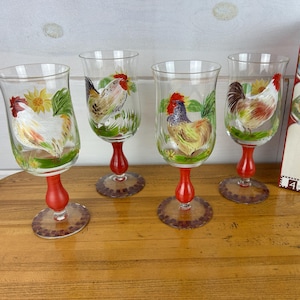 May include: Set of four clear wine glasses with hand-painted rooster designs. Each glass features a different rooster illustration with red, yellow, and green accents. The glasses have red stems and decorative bases, with a matching box.