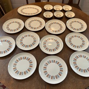 May include: A set of 13 white ceramic dinnerware plates with a blue and orange geometric pattern. The plates are arranged on a wooden table.