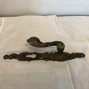 May include: Ornate brass door handle with a keyhole and a decorative design. The handle is on a white fabric background.