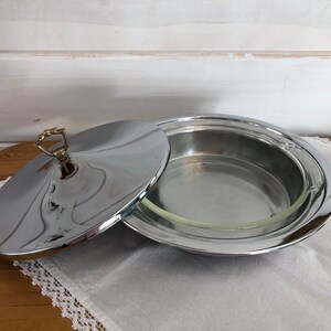 May include: A silver serving dish with a glass insert and a lid with a gold knob. The dish is round and has a raised edge.