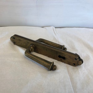 May include: Antique brass door handle with a keyhole and two long levers. The handle is on a white fabric surface.