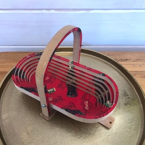 May include: A red and white wooden basket with a Coca-Cola design. The basket has a handle and is sitting on a gold tray.