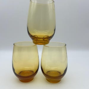 May include: Three amber-colored stemless glasses. The glasses have a rounded shape with a slightly flared rim and a thick, amber-colored base. The glasses are arranged in a pyramid shape against a white background.