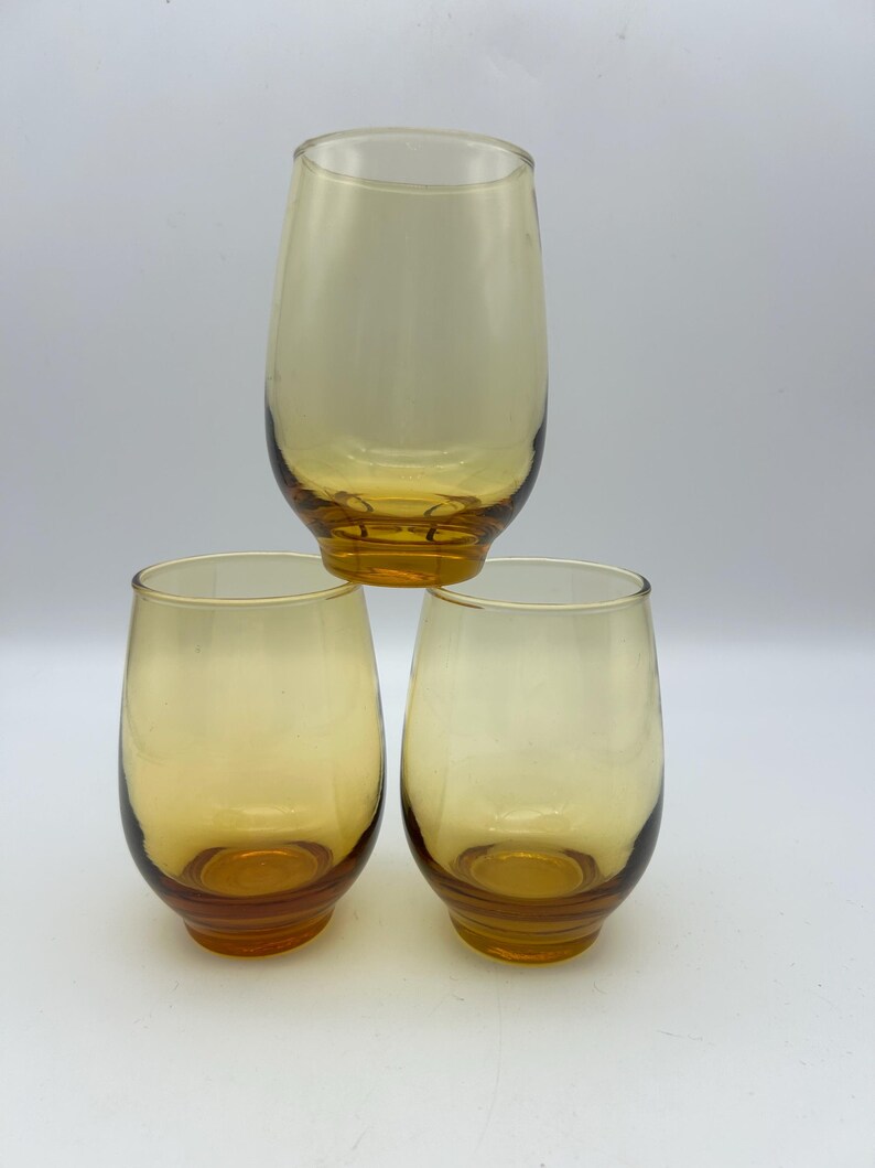May include: Three amber-colored stemless glasses. The glasses have a rounded shape with a slightly flared rim and a thick, amber-colored base. The glasses are arranged in a pyramid shape against a white background.