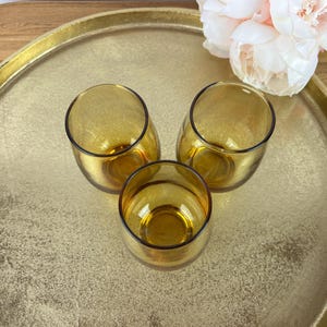 May include: Three amber-colored, stemless drinking glasses with a rounded shape are arranged on a gold-colored tray. The glasses are empty and have a dark rim. A bouquet of pink flowers is in the background.