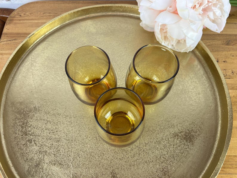 May include: Three amber-colored, stemless drinking glasses with a rounded shape are arranged on a gold-colored tray. The glasses are empty and have a dark rim. A bouquet of pink flowers is in the background.