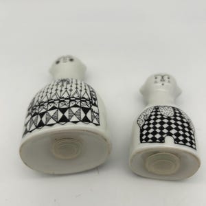 May include: A pair of ceramic salt and pepper shakers in the shape of people. The shakers are white with black geometric patterns. The salt shaker has a black and white checkered pattern and the pepper shaker has a black and white geometric pattern.