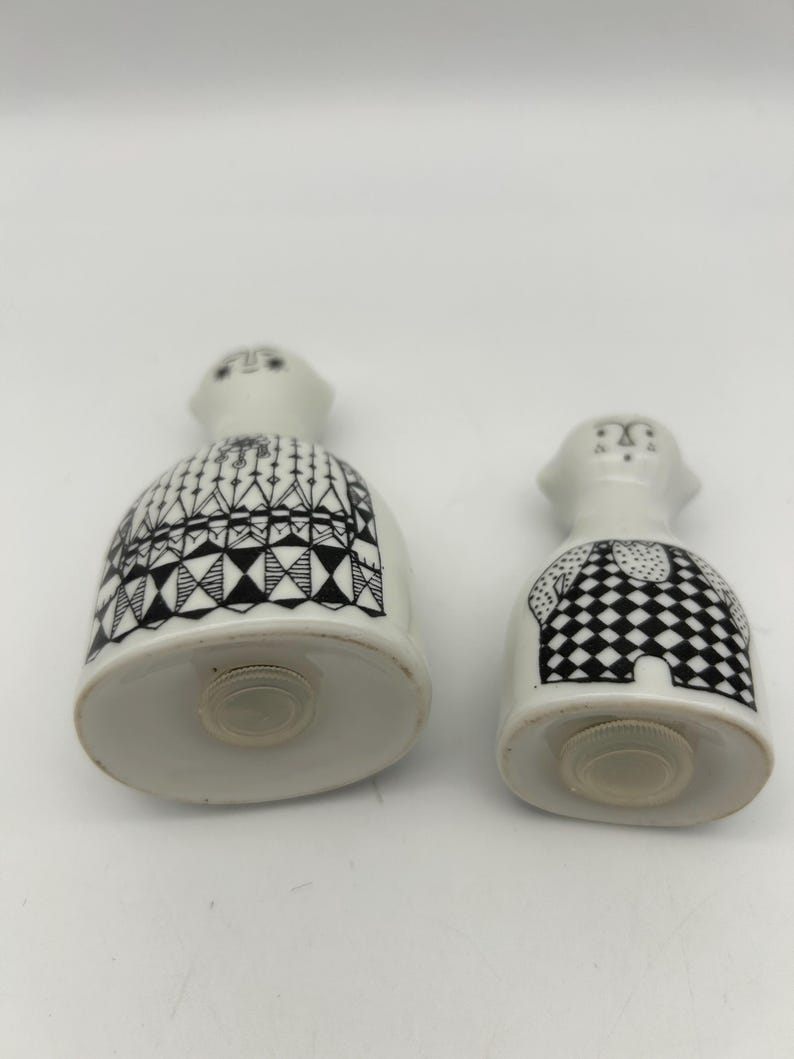 May include: A pair of ceramic salt and pepper shakers in the shape of people. The shakers are white with black geometric patterns. The salt shaker has a black and white checkered pattern and the pepper shaker has a black and white geometric pattern.