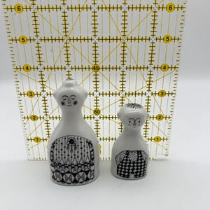 May include: A pair of ceramic salt and pepper shakers in the shape of people. The shakers are white with black and white geometric patterns. The female shaker has a long dress and a necklace. The male shaker has a short-sleeved shirt and pants.