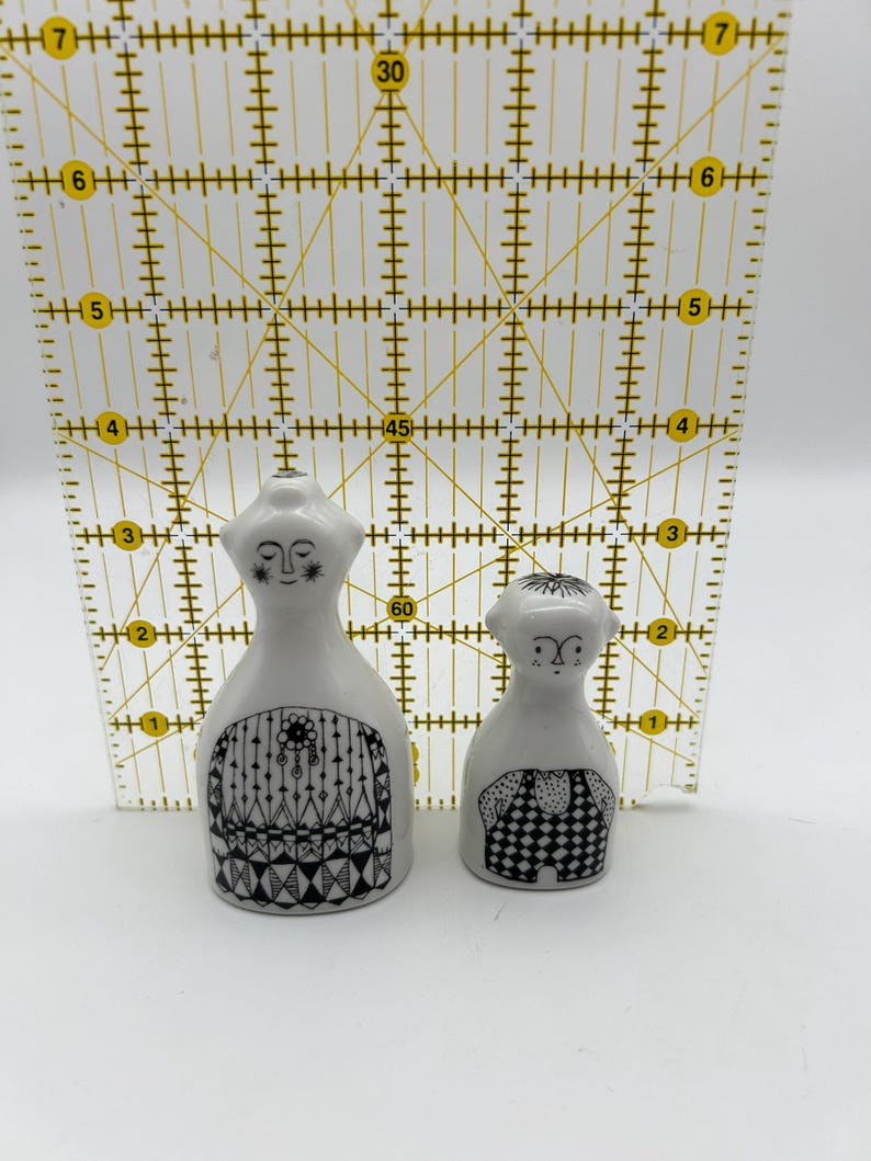 May include: A pair of ceramic salt and pepper shakers in the shape of people. The shakers are white with black and white geometric patterns. The female shaker has a long dress and a necklace. The male shaker has a short-sleeved shirt and pants.