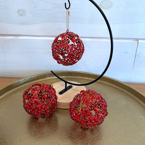 May include: Three red jeweled Christmas ornaments on a gold tray with a black metal stand. The ornaments are covered in red rhinestones.