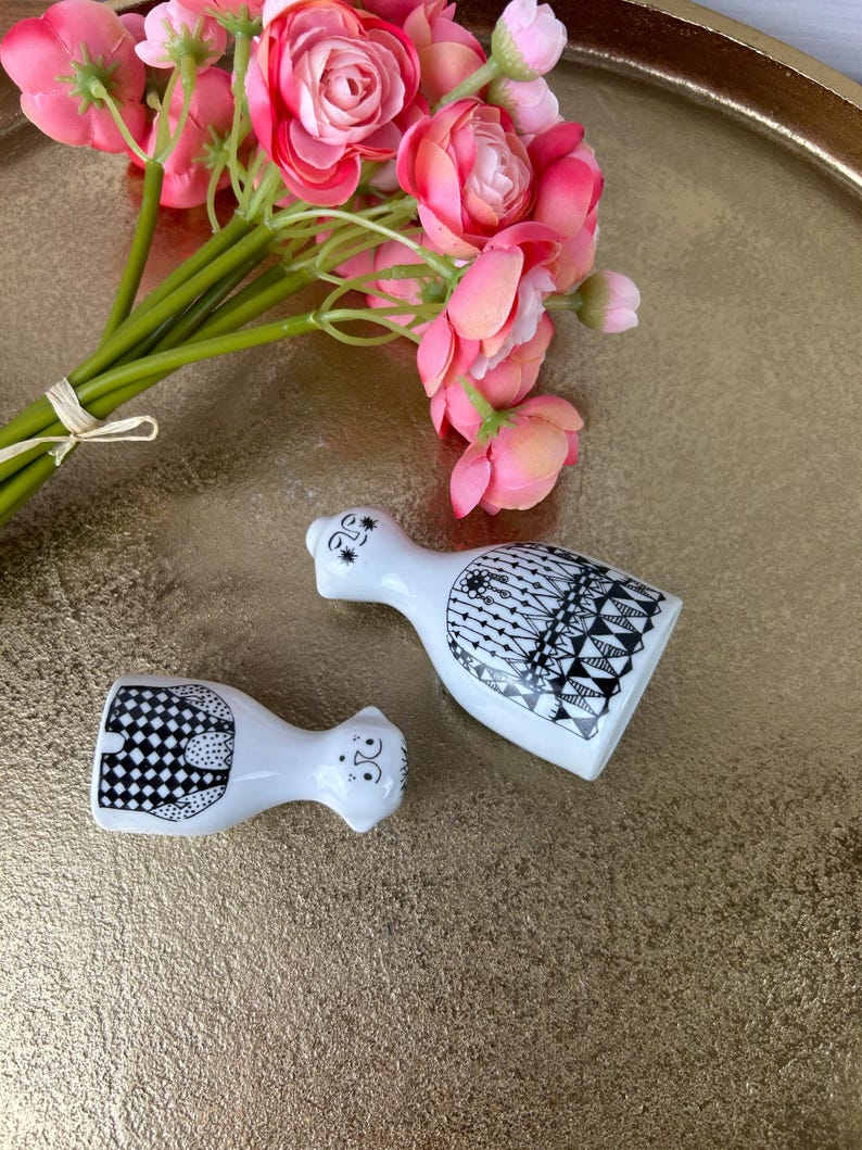 May include: A pair of white ceramic salt and pepper shakers with black and white geometric designs. The shakers are shaped like people and have faces.