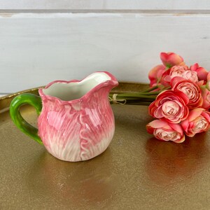 May include: A pink ceramic creamer in the shape of a flower with a green handle. The creamer is sitting on a gold tray with a bouquet of pink flowers.
