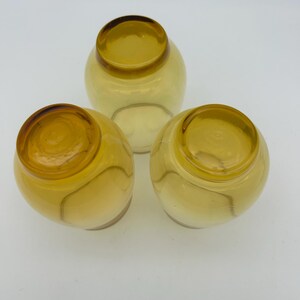 May include: Three amber-colored, round glass bowls. Each bowl has a rounded base and a slightly flared rim. The glass has a warm, golden hue, and the bowls are arranged in a triangular formation.