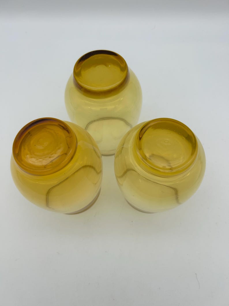 May include: Three amber-colored, round glass bowls. Each bowl has a rounded base and a slightly flared rim. The glass has a warm, golden hue, and the bowls are arranged in a triangular formation.