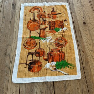 May include: A rectangular kitchen towel featuring a vintage illustration of copper cookware, vegetables, and utensils against a wood-grain background. The towel has a white border and is made of a light-colored fabric.