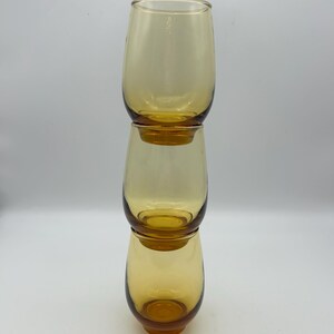 May include: Three amber-colored, stemless drinking glasses stacked vertically. The glasses have a rounded shape and a smooth, transparent surface. The glasses are made of glass and are empty.
