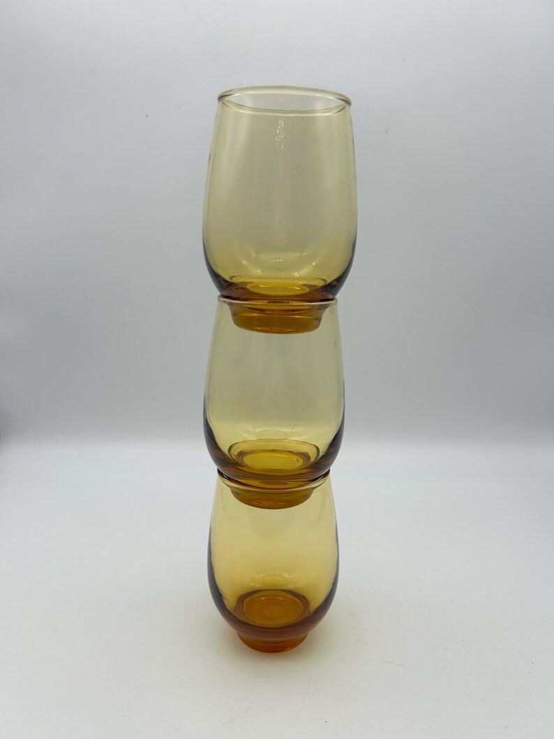 May include: Three amber-colored, stemless drinking glasses stacked vertically. The glasses have a rounded shape and a smooth, transparent surface. The glasses are made of glass and are empty.