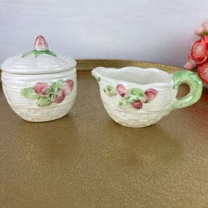 May include: A vintage cream-colored ceramic sugar bowl with a lid and a creamer. Both pieces are decorated with pink and green floral accents and a woven basket design. The creamer has a green handle.