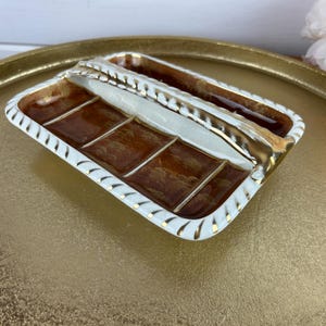May include: A rectangular, vintage butter dish with a brown interior and a white and gold rim. The dish has a central divider and a gold handle. The dish is placed on a gold-colored tray.