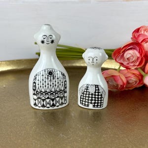 May include: A pair of white ceramic salt and pepper shakers with black and white geometric designs. The shakers are shaped like people with faces and are standing on a gold tray.
