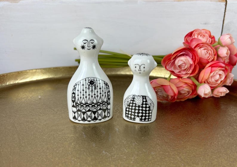May include: A pair of white ceramic salt and pepper shakers with black and white geometric designs. The shakers are shaped like people with faces and are standing on a gold tray.