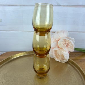May include: Three amber-colored, stemless drinking glasses stacked on top of each other. The glasses are on a gold-colored tray, with a bouquet of light pink flowers in the background. The glasses have a rounded shape.