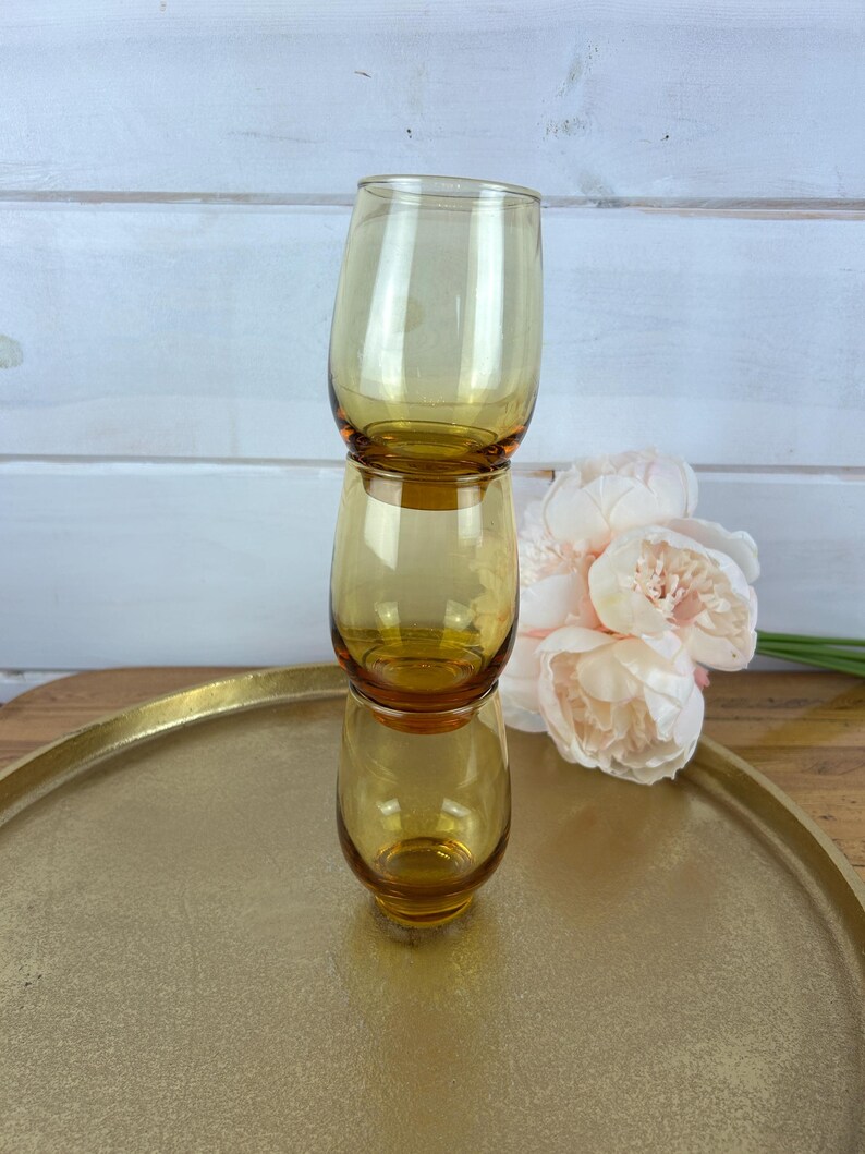 May include: Three amber-colored, stemless drinking glasses stacked on top of each other. The glasses are on a gold-colored tray, with a bouquet of light pink flowers in the background. The glasses have a rounded shape.