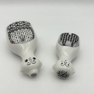 May include: Two white ceramic figurines with black and white designs. The figurines are shaped like people and have simple faces.