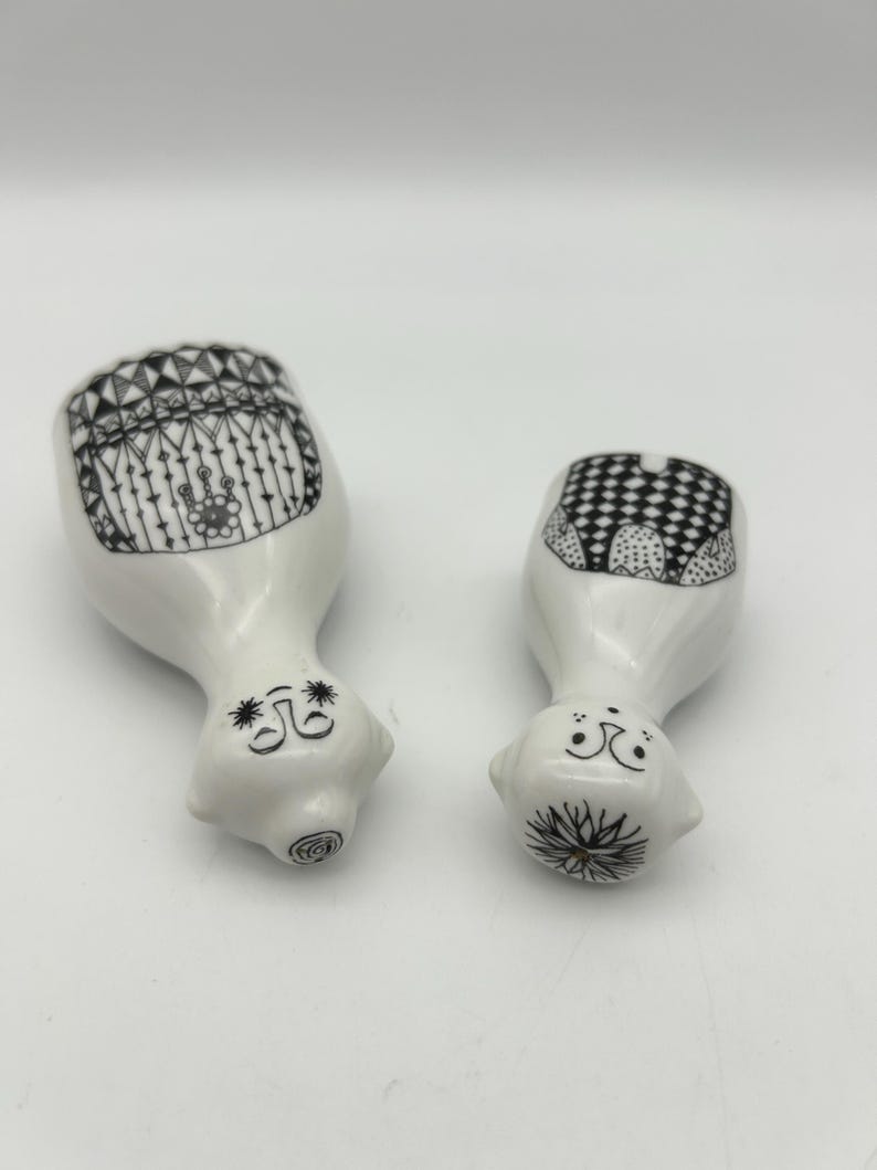 May include: Two white ceramic figurines with black and white designs. The figurines are shaped like people and have simple faces.