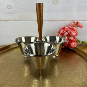 May include: A vintage condiment server with three stainless steel bowls and a wooden handle. The server sits on a gold-colored tray, with a bouquet of pink flowers in the background. The bowls are approximately 3 inches in diameter.