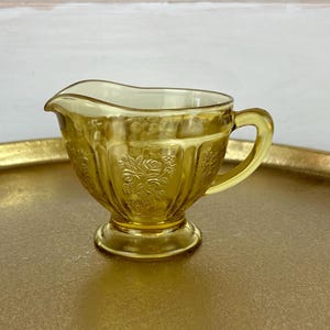 May include: A small, yellow glass creamer with a floral design. The creamer has a handle and a round base.