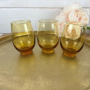 May include: Three amber-colored, stemless drinking glasses with a rounded base and tilted design. The glasses sit on a gold-colored tray. A bouquet of light pink flowers is in the background.