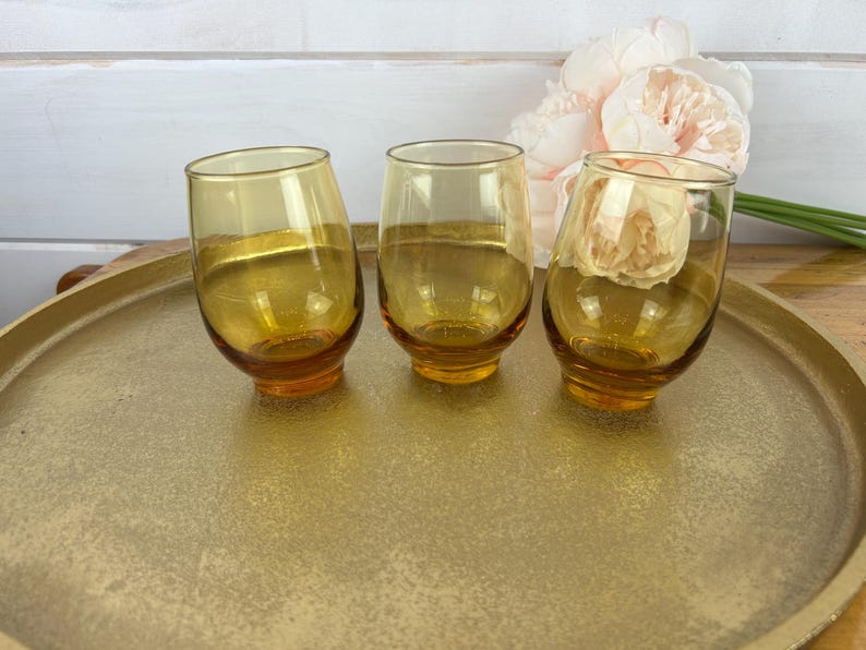 May include: Three amber-colored, stemless drinking glasses with a rounded base and tilted design. The glasses sit on a gold-colored tray. A bouquet of light pink flowers is in the background.