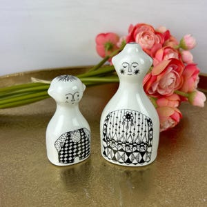 May include: Two white ceramic figurines with black and white geometric patterns. The figurine on the left is a person with a star on their head and a checkered pattern on their body. The figurine on the right is a person with a star on their forehead and a geometric pattern on their body.