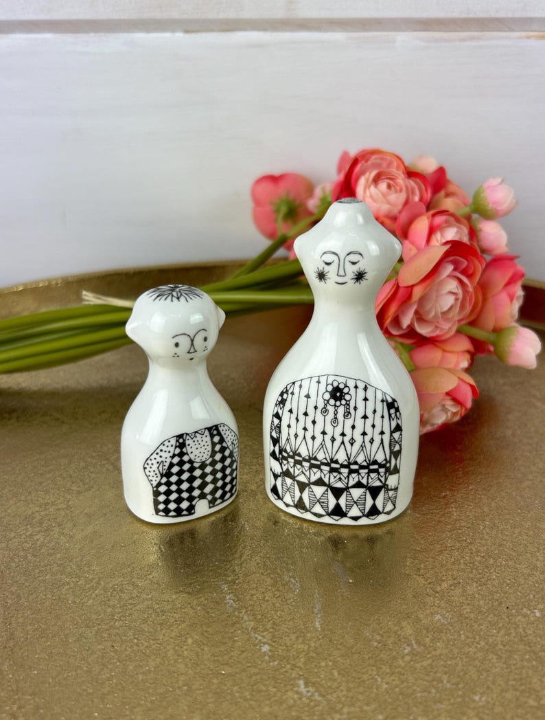 May include: Two white ceramic figurines with black and white geometric patterns. The figurine on the left is a person with a star on their head and a checkered pattern on their body. The figurine on the right is a person with a star on their forehead and a geometric pattern on their body.